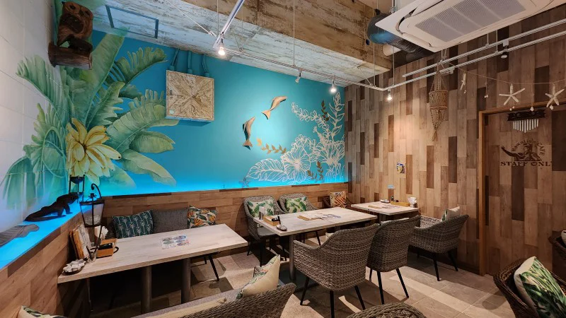 Dining area with tropical wall art and wicker chairs at Hawaiian Café Kapuka in American Village, Chatan