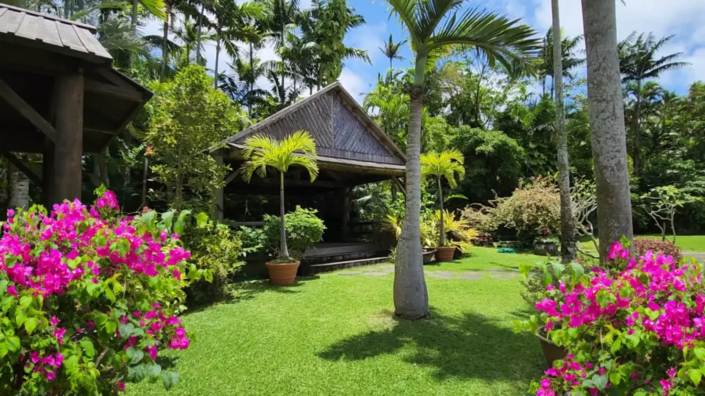 Wooden pavilion surrounded by palm trees, tropical plants, and pink bougainvillea flowers at Southeast Botanical Gardens in Okinawa, Japan.