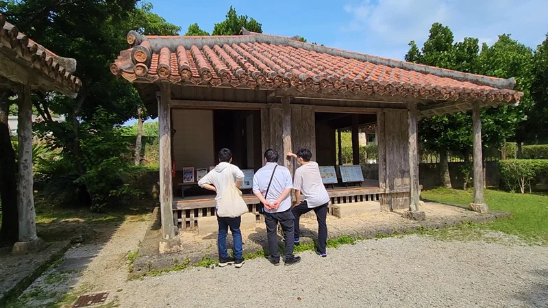 Visitors observing a traditional Okinawan wooden house with red-tiled roof.