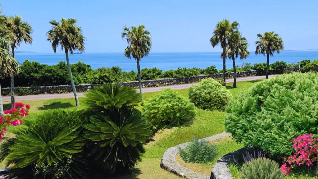 Tropical garden at Ocean Expo Park in Okinawa with palm trees, lush green plants and a bright blue ocean in the background under a clear sky.