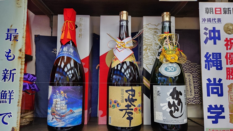 Three large bottles of premium Awamori with colorful, ornate packaging displayed on a shelf.
