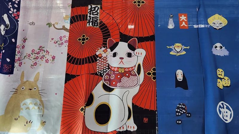 Noren curtains decorated with Totoro, a lucky cat, and other Japanese motifs displayed in a shop.