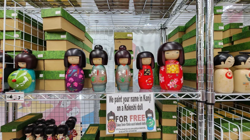 Shelves of traditional Japanese Kokeshi dolls with a sign offering custom Kanji name painting.