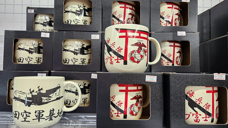 Stacked souvenir mugs with Okinawa marine and military-themed illustrations displayed in a shop.