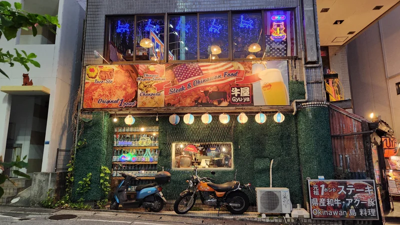 Nighttime view of a restaurant in Okinawa serving steak and local dishes, with lanterns and motorbikes outside.