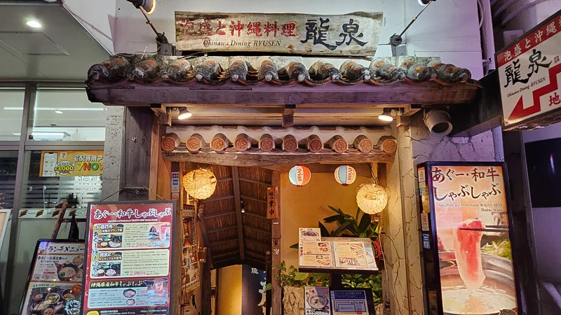 Traditional Okinawan restaurant entrance with tiled roof, lanterns, and menu boards at night.