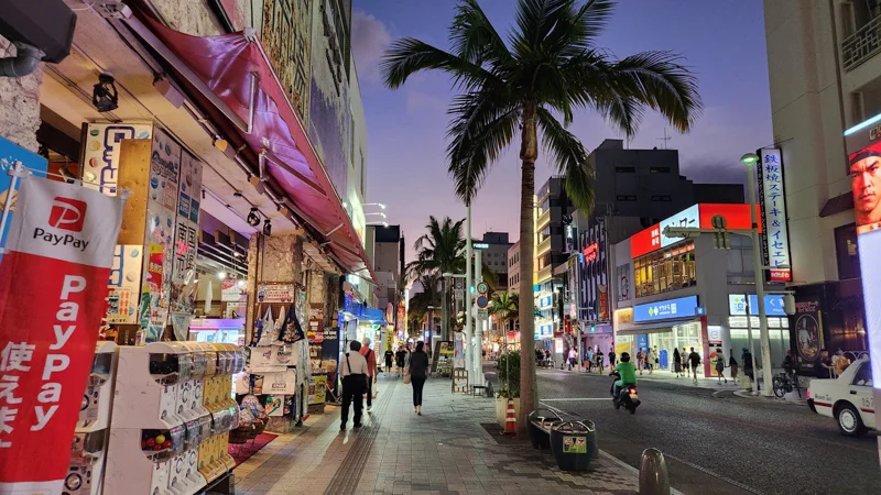 Pedestrians walking along Kokusai Dori in Naha at dusk, with lit storefronts and palm trees lining the street.