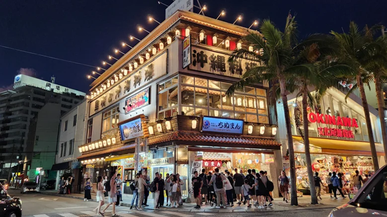 Crowded nighttime scene outside a multi-story Okinawan restaurant with bright lanterns and tropical palm trees.