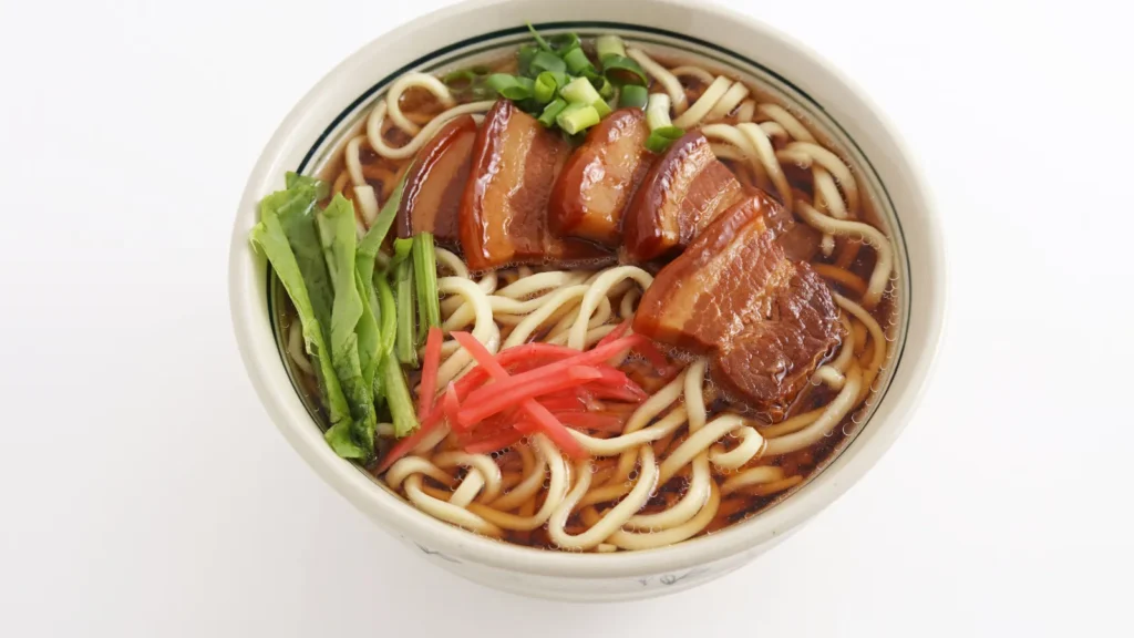 Bowl of Okinawa soba topped with braised pork belly, green onions, greens, and pickled ginger