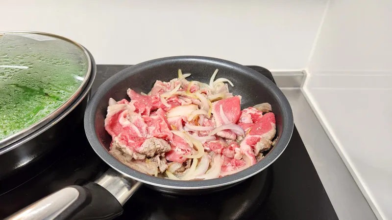 Thinly sliced beef and onions cooking together in a frying pan on a stovetop