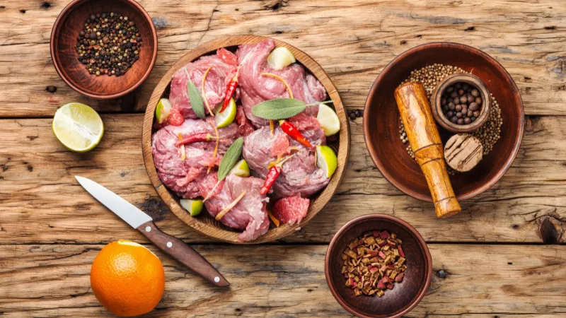 Morceaux de viande crue marinés avec des agrumes, des herbes et des épices sur une table en bois.
