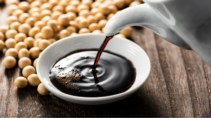 Soy sauce being poured from a container into a white bowl with soybeans nearby