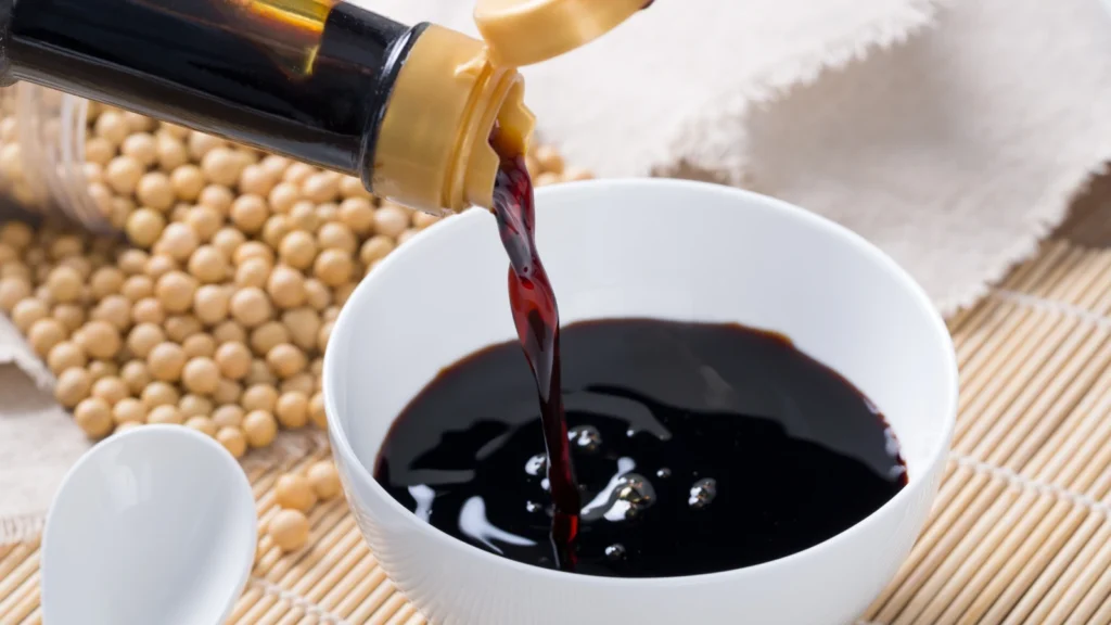 Soy sauce being poured from a bottle into a white bowl with soybeans in the background