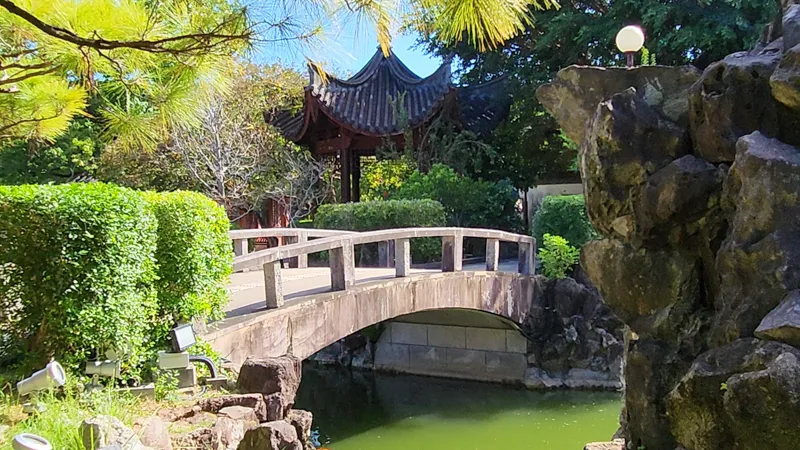Arched stone bridge over green water with a pavilion in the background at Fukushuen Garden